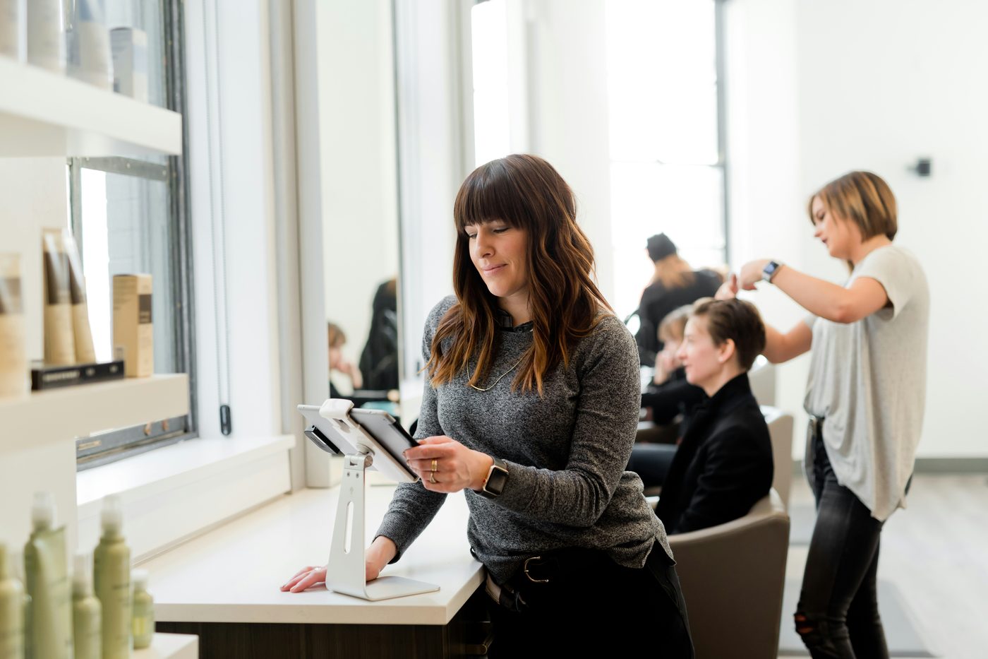 A salon owner taking a booking on a point-of-sale tablet while stylists work behind her